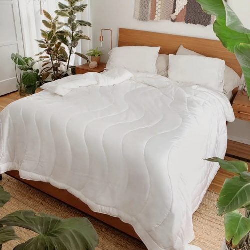 A white, wavy-patterned comforter and matching pillows are neatly arranged on a wooden bed frame, surrounded by lush potted plants and warm wooden accents.