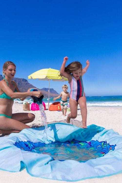 A woman is filling a small inflatable pool with water while two children play joyfully on the beach under a sunny sky.