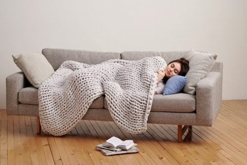 A young woman sleeps peacefully on a grey sofa, cozying up under a thick, light grey chunky knit weighted blanket.