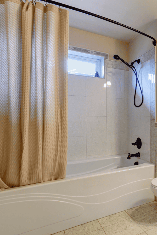 Beige fabric shower curtain hanging from a curved rod over a white bathtub with tiled walls and a bronze showerhead.