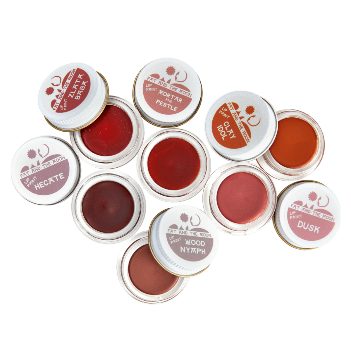 An overhead flat-lay displays nine small open pots of natural tinted lip balm or cheek stain from Fat and The Moon in shades of reddish-pink and brown, with their labeled white lids scattered around them on a black background.