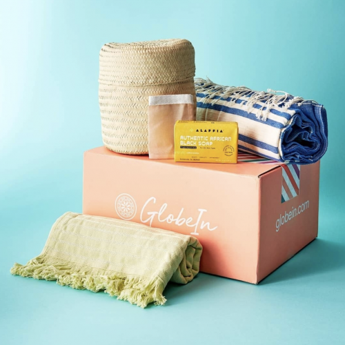 A large peach-colored GlobeIn subscription box is shown against a blue background, topped with a straw basket, a bar of Alaffia African Black Soap, a rolled blue and white striped towel, and a folded light green fringed towel.