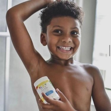 A young child with dark skin and short dark hair is smiling and looking at the camera while applying a Play Pits natural deodorant stick to their underarm.
