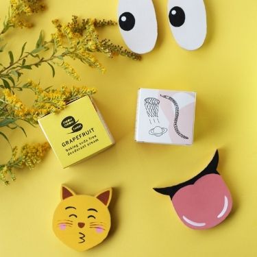 A flatlay of Meow Meow Tweet's grapefruit scented baking soda-free deodorant cream with emjoi cutouts and yellow flowers on a yellow background.