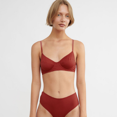 A white feminine presenting person with short blonde hair stands looking at the camera in an eco-friendly red underwired bra and panty from Araks