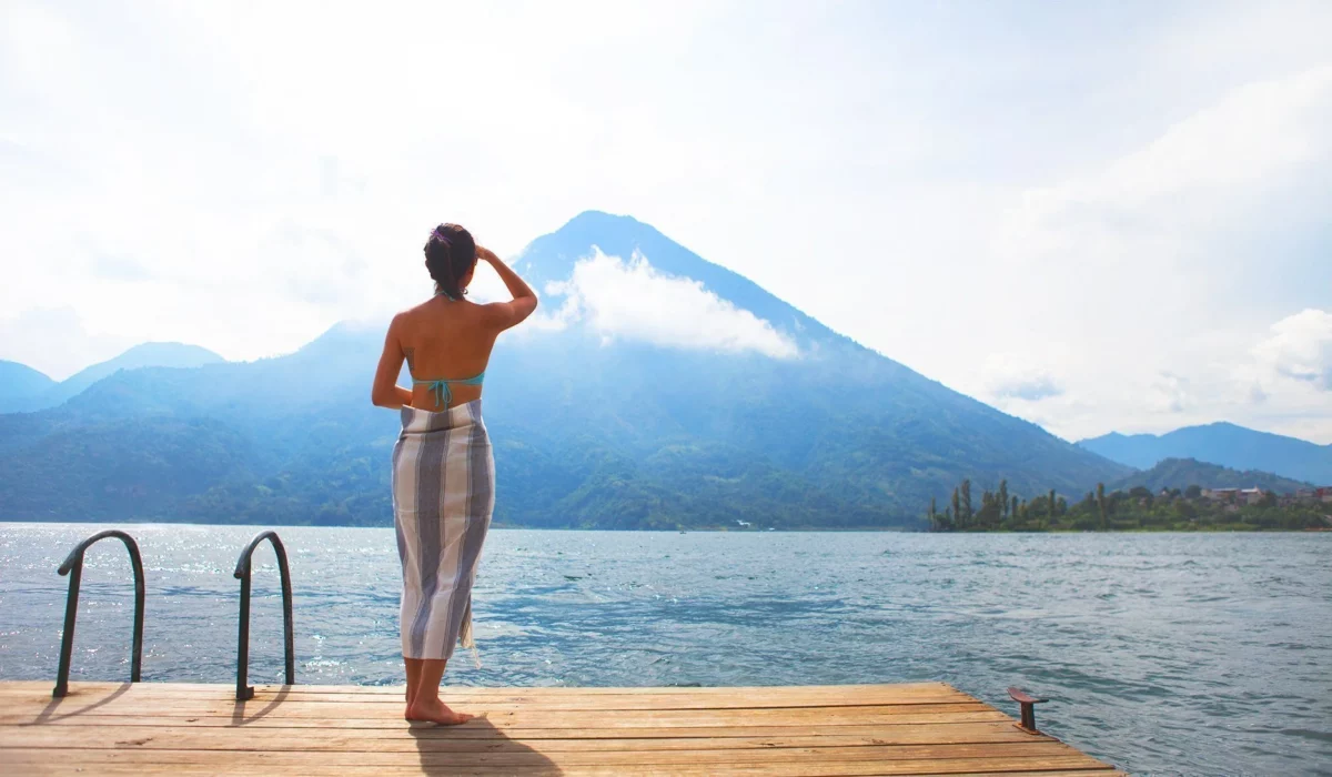 A woman standing on the beach, wrapped in a Stray and Wander organic beach towel, with a stunning mountain view in the background.
