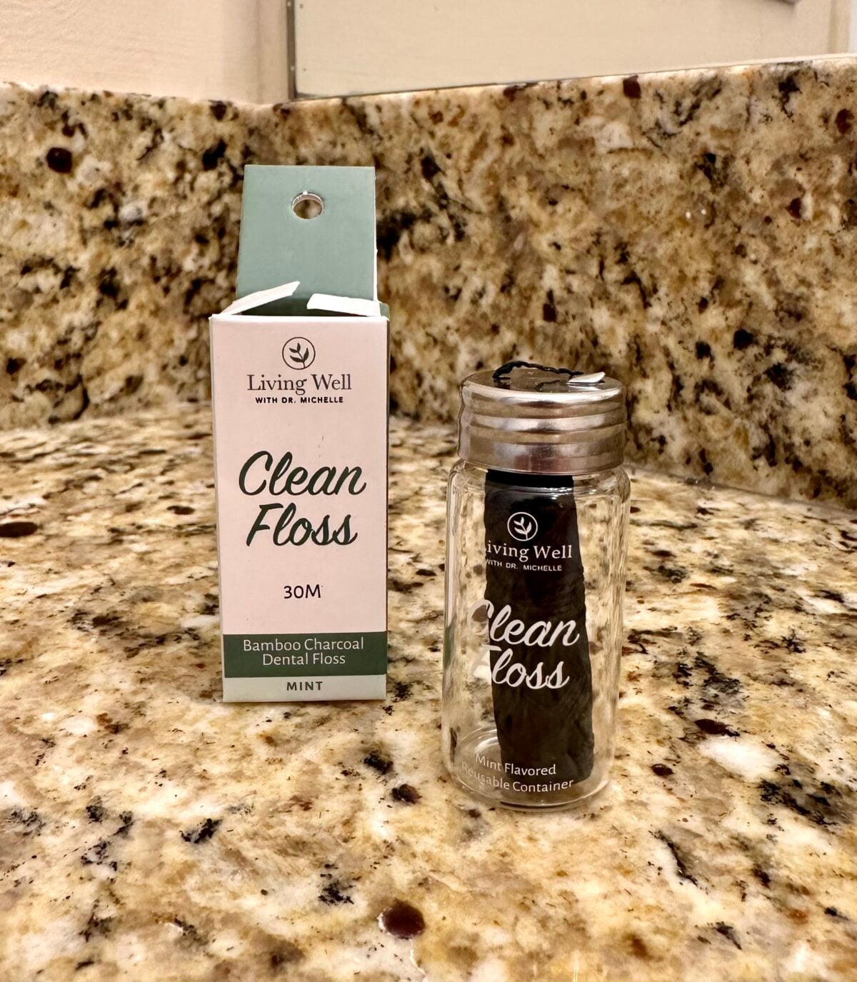 Living Well &lsquo;Clean Floss&rsquo; box and a refillable glass jar holding black bamboo charcoal dental floss displayed on a granite countertop.