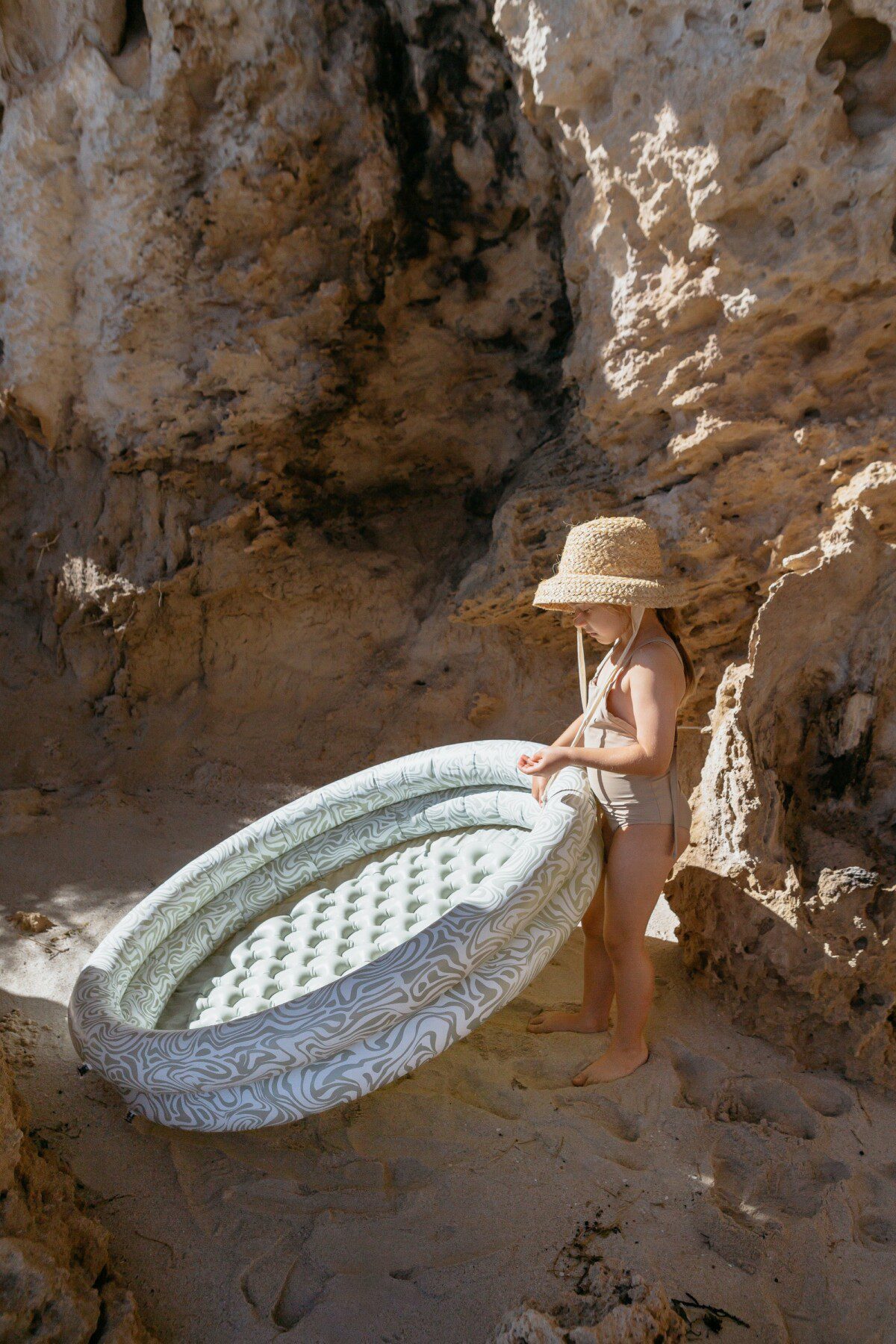 The photo shows a child holding a green and white patterned inflatable pool, standing on sandy ground with rocky cliffs in the background. The child is wearing a straw hat and a swimsuit.