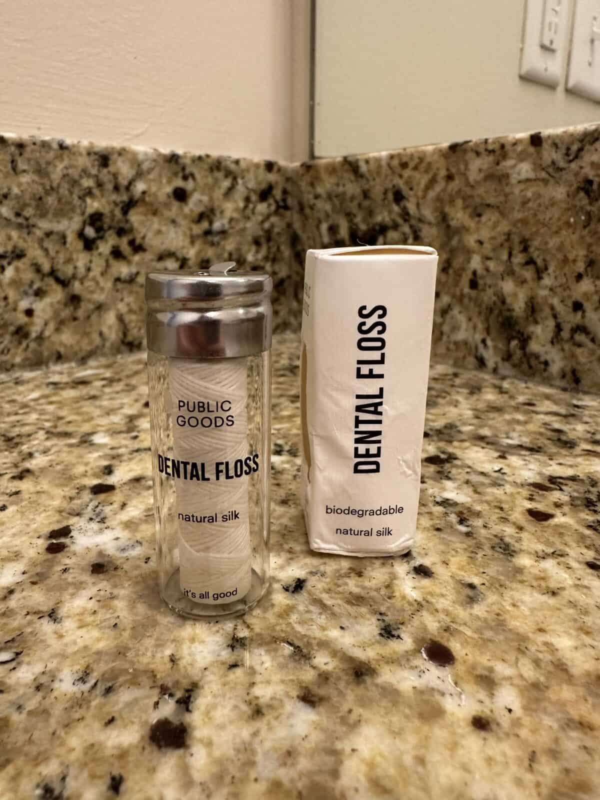 Glass container of Public Goods natural silk dental floss next to its white biodegradable paper box on a granite countertop.