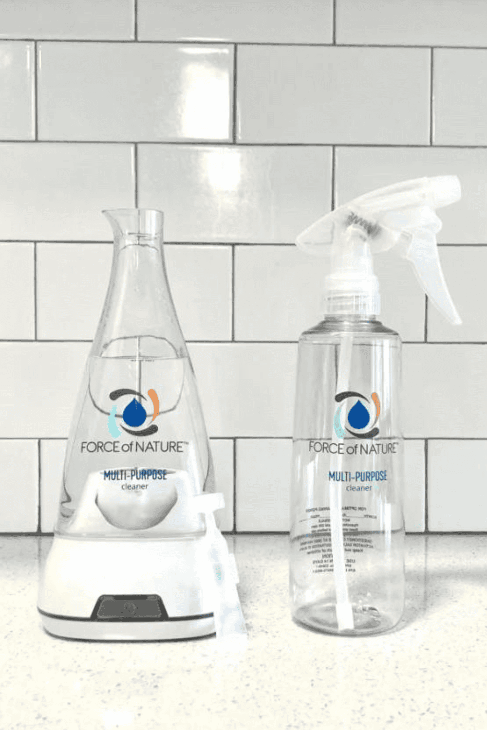 A shot focuses on two clear bottles of Force of Nature Multi-Purpose Cleaner, one being created in the proprietary electrolysis flask and the other a ready-to-use spray bottle, set against a white subway tile backsplash.