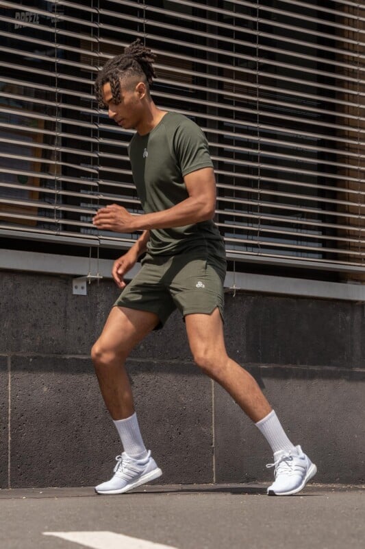 A dark-haired man with dreadlocks is running on the sidewalk in a dark olive green athletic shirt and shorts from Boldwill, paired with white socks and sneakers, in front of a modern building facade.