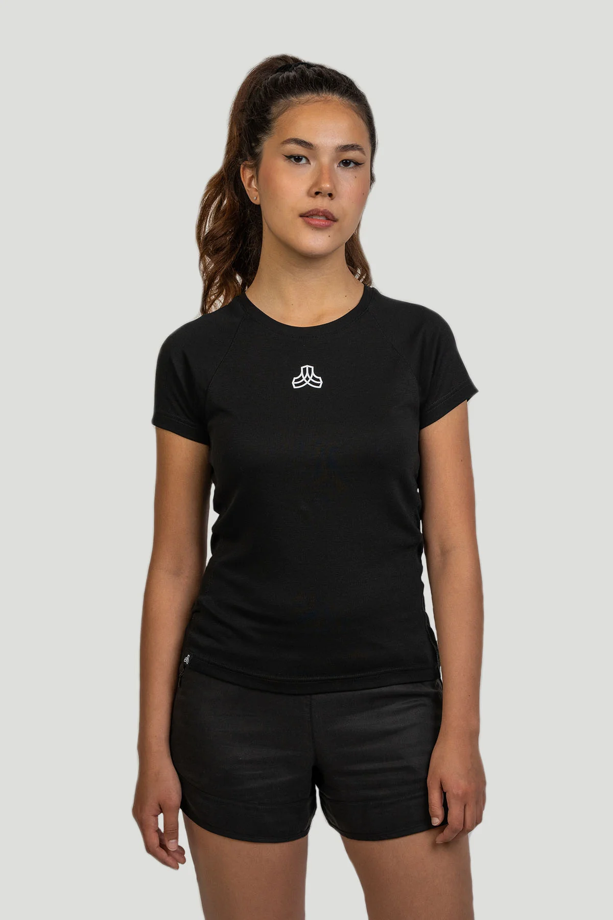 A female model with her hair in a high ponytail is standing straight and looking forward, wearing a fitted black short-sleeve athletic top with a small white logo on the chest and dark shorts from Boldwill.