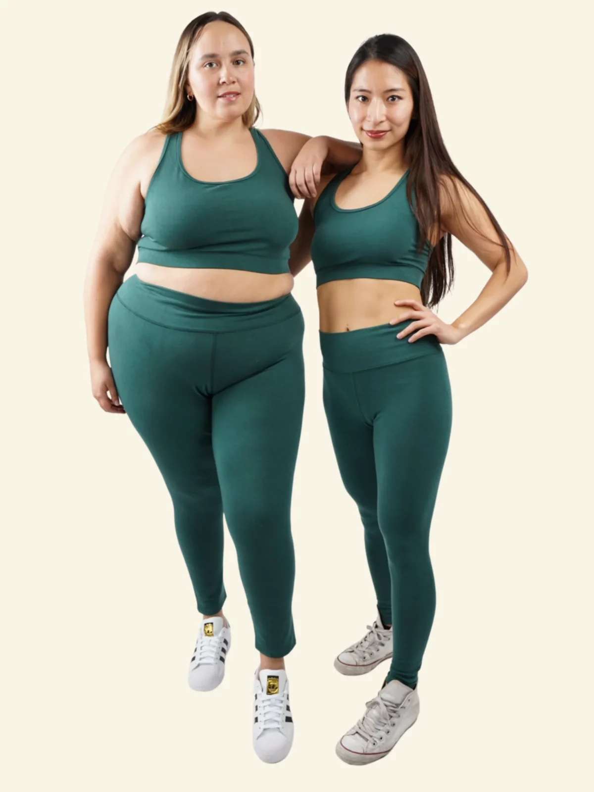 Two women of different body types and ethnicities are standing together, smiling, and wearing matching dark green sports bras and high-waisted leggings from Reprise, paired with white sneakers.