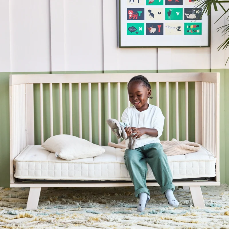 A young child sits on a wooden daybed, smiling while putting on a sock. The bed has a mattress and pillow, and a colorful animal-themed art print hangs on the wall above.