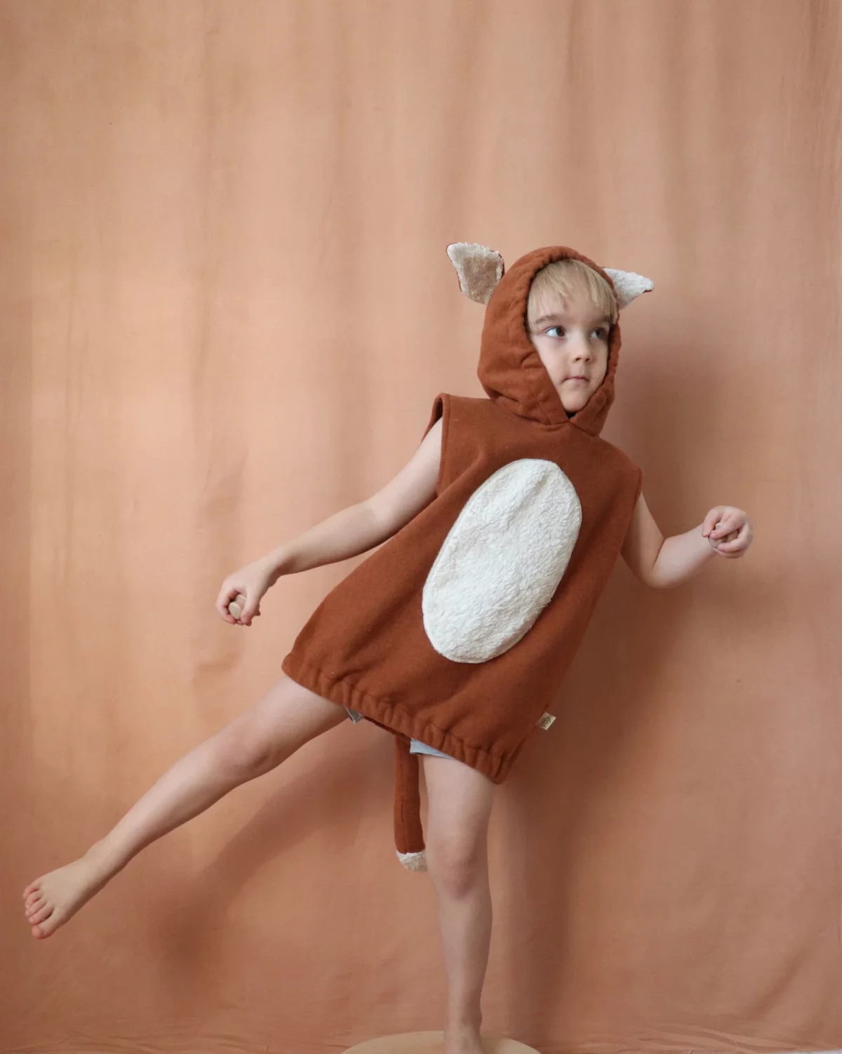 A barefoot child in a sleeveless, rust-colored fox costume with a hood, ears, and a cream-colored belly patch is standing on one leg against a beige background.