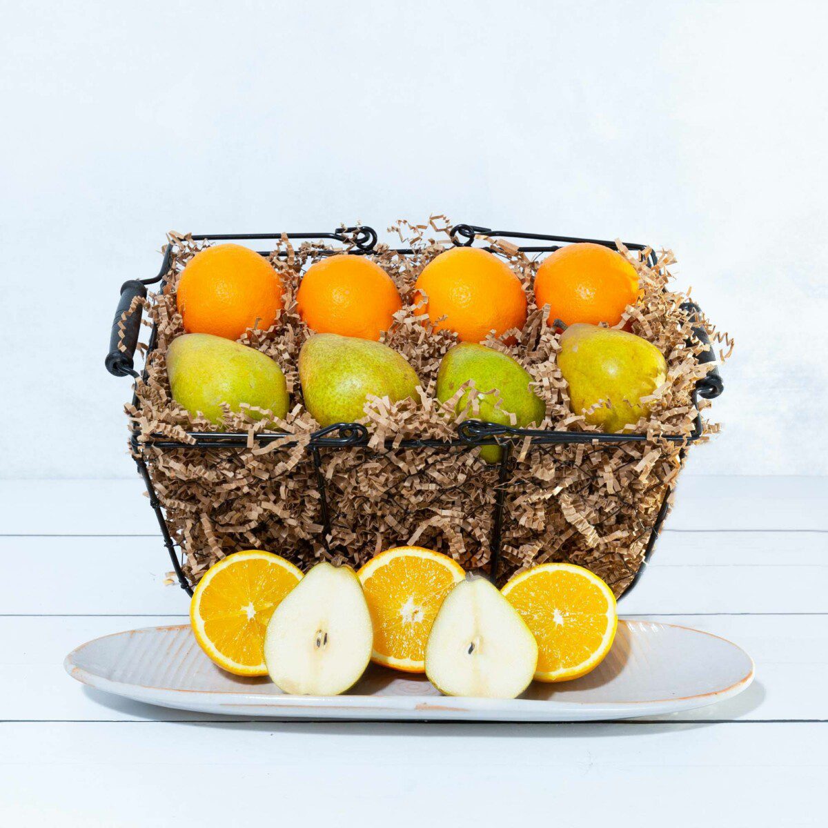 
A studio shot features a metal wire basket filled with four oranges and four green pears over paper shred filler, with an arrangement of sliced oranges and pears on a long platter in the foreground.