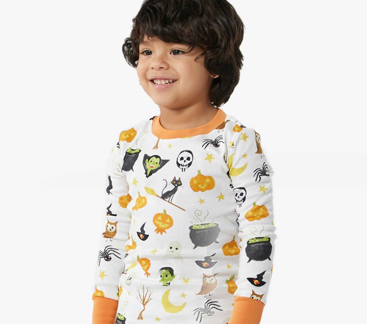 A happy young boy with a wide smile is wearing a white pajama set from Pottery Barn Kids with a colorful, all-over pattern of Halloween-themed images like pumpkins, spiders, and witches' cauldrons.