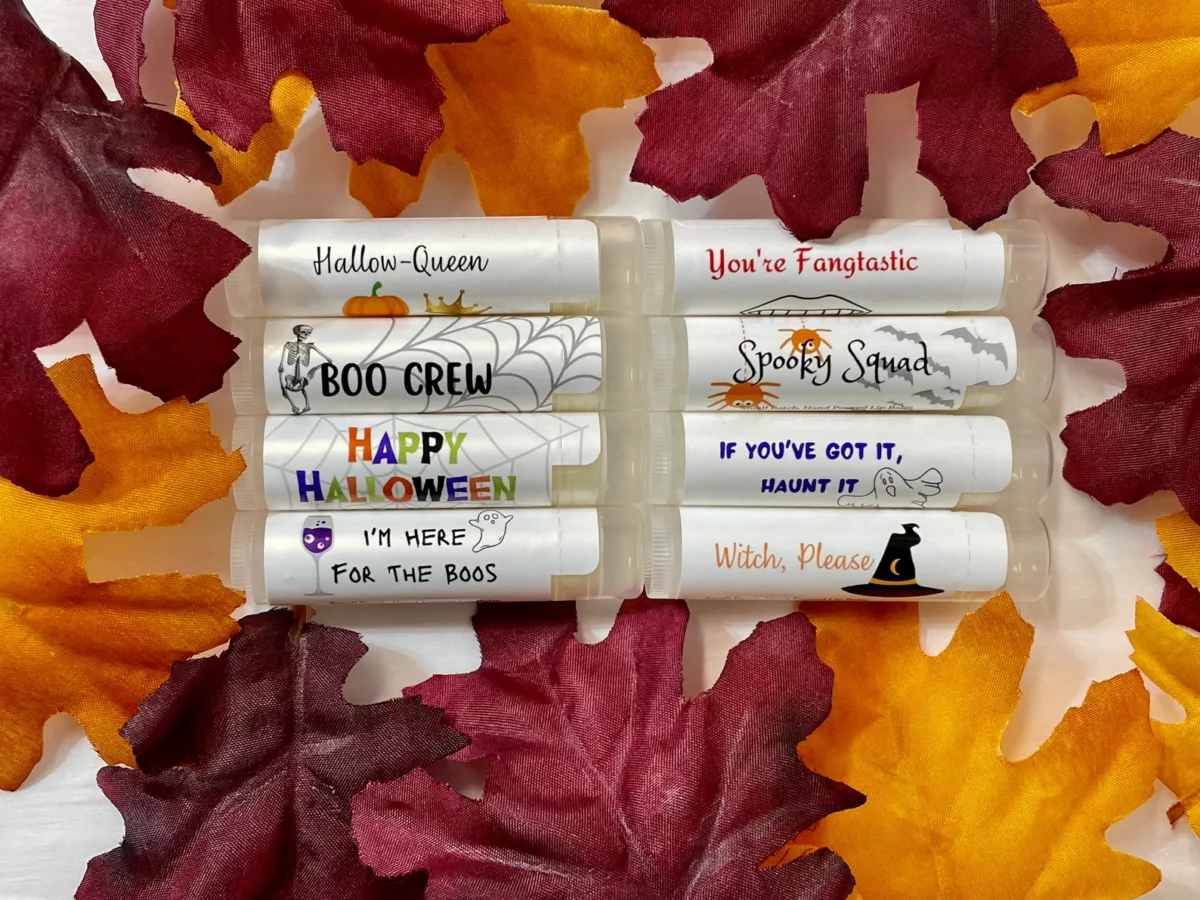 Eight lip balm tubes with Halloween-themed labels are arranged in two columns on a white surface and bordered by artificial autumn leaves in shades of red, burgundy, and orange.