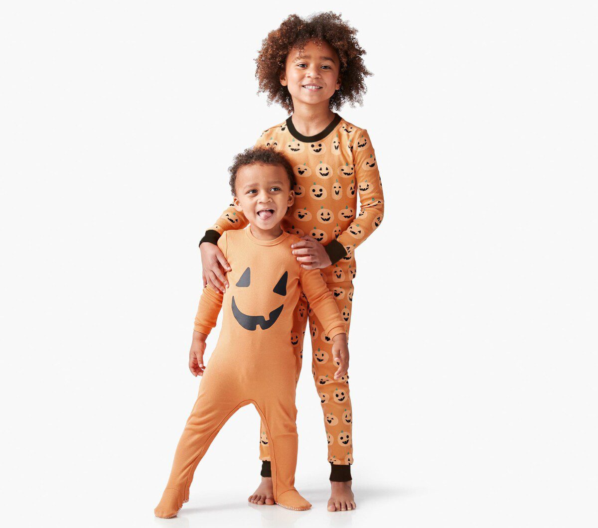 Two smiling young children, one wearing a solid orange pumpkin-face onesie and the other in matching pumpkin-patterned pajamas from Pottery Barn Kids, are standing together on a white background.