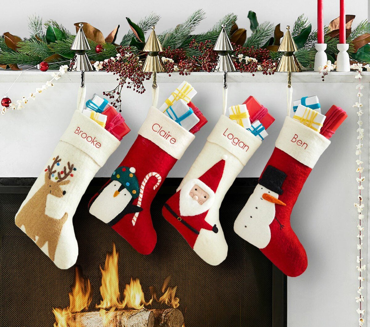Four felt Christmas stockings—personalized with the names Brooke, Claire, Logan, and Ben and featuring a reindeer, a penguin, Santa, and a snowman—are filled with gifts and hung above a burning fireplace.