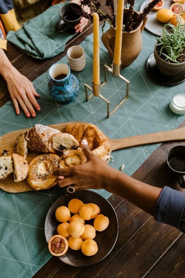 an organic table runner, and placemats arranged on a wooden dining table with two people having a coffee and getting bread