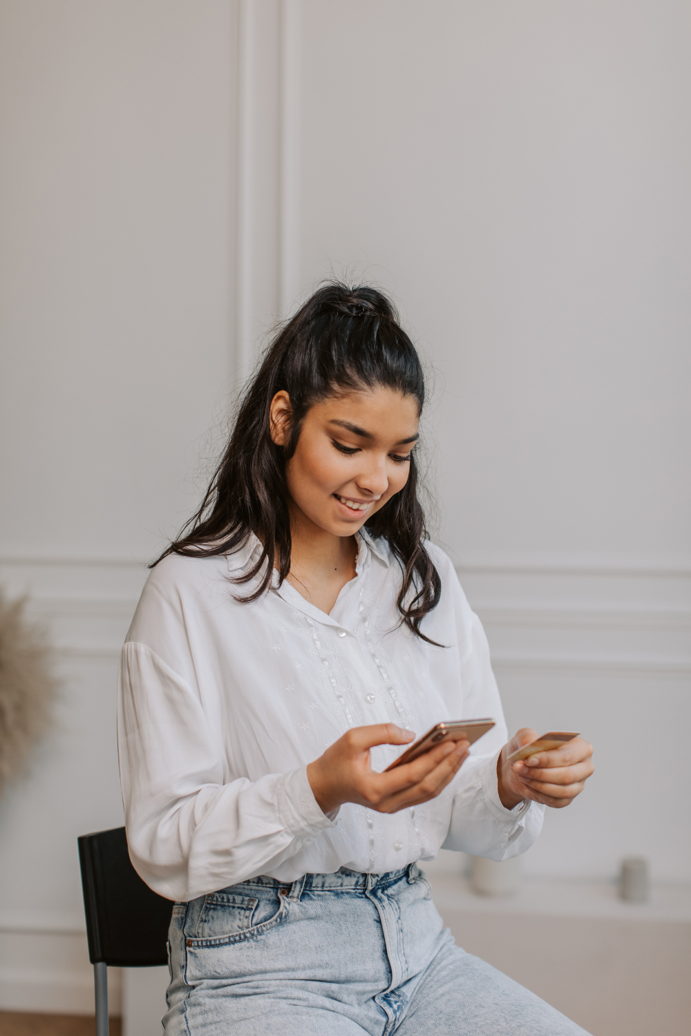 A young woman with dark hair in a half-up ponytail is smiling and looking down at a smartphone held in one hand and a credit card held in the other, wearing a white blouse and light wash jeans against a soft, light-colored background.