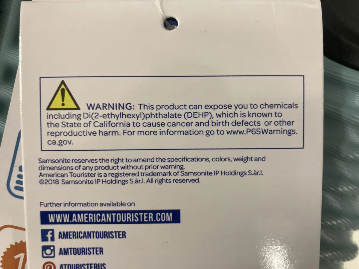 The photo shows a warning tag from American Tourister, stating that the product can expose users to chemicals, including Di(2-ethylhexyl)phthalate (DEHP), which is known to the state of California to cause cancer, birth defects, or other reproductive harm.