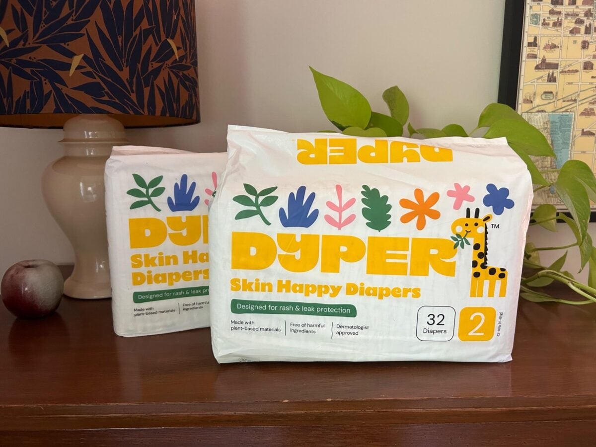 Two packages of Dyper Skin Happy Diapers, size 2, sit on a wooden surface. The packaging features colorful plants and a giraffe illustration. A lamp, apple, and leafy plant are in the background.