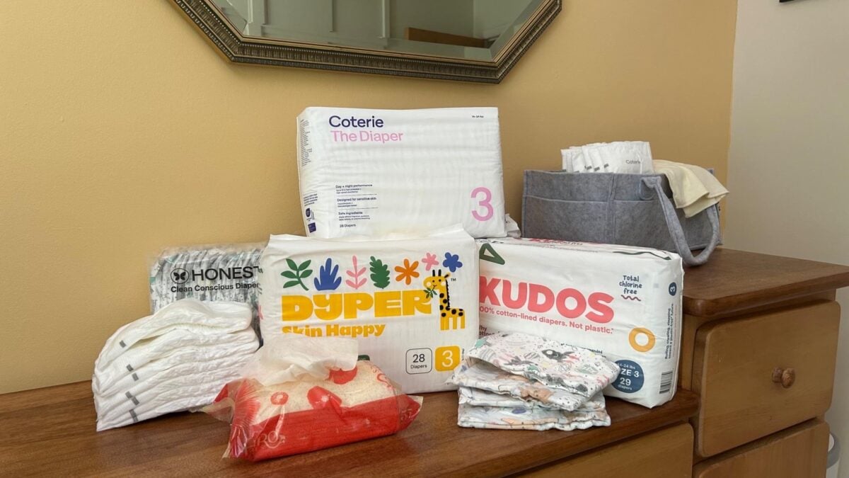 A wooden dresser holds packs of baby diapers from Coterie, Kudos, Dyper, and Honest, with some diapers stacked loosely nearby and a gray storage bin containing more diapers in the background.