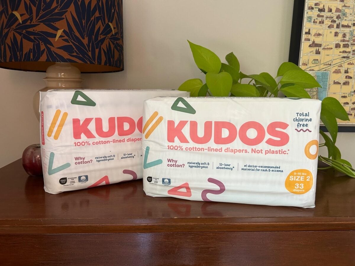 Two packs of Kudos size 2 cotton-lined diapers rest on a wooden surface near a potted plant, a lamp with a patterned shade, and a framed map. The packaging states 100% cotton-lined diapers. Not plastic..