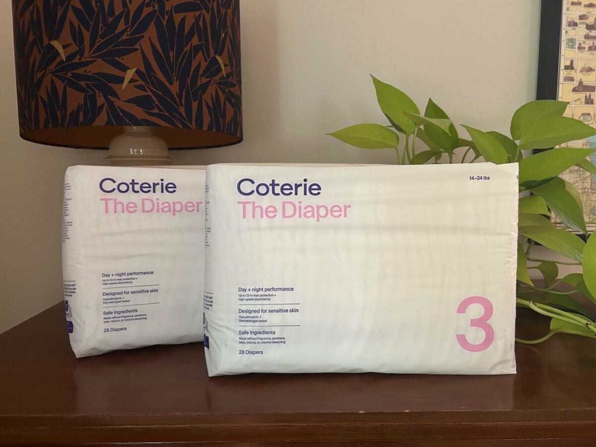 Two packages of Coterie The Diaper, each labeled with a large pink number 3, sit on a wooden surface near a leafy plant and a lamp with a dark patterned shade.