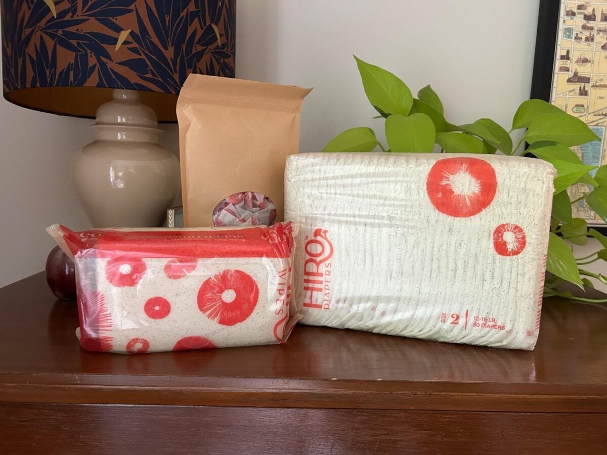 Two packs of diapers with red and white designs and a brown paper bag are displayed on a wooden surface. A leafy plant, a lamp with a patterned shade, and a framed map are in the background.