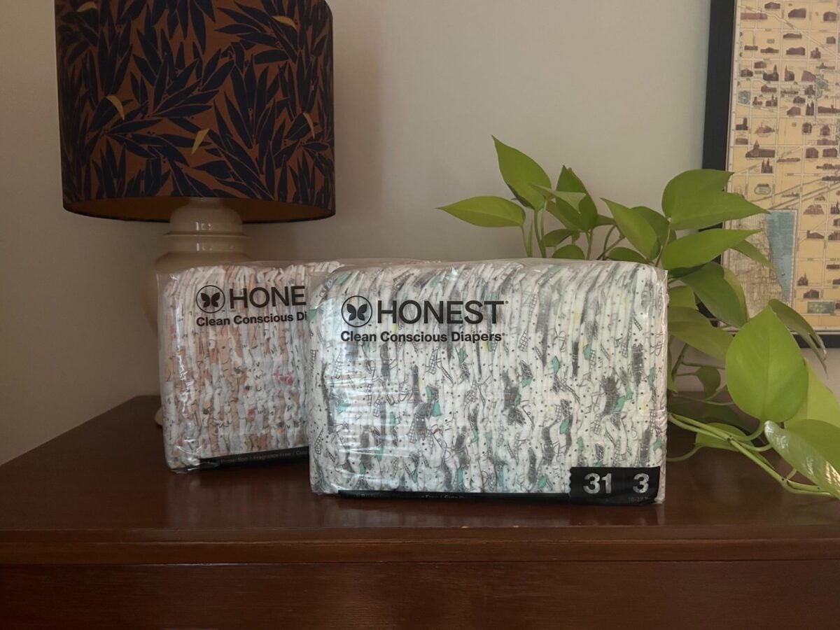 Two packages of Honest brand Clean Conscious Diapers are placed on a wooden surface next to a green leafy plant and a lamp with a patterned shade. The packaging shows a size of 3 and a quantity of 31 diapers.
