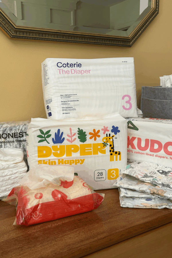 Various brands of size 3 diapers, including Coterie, Dyper, Kudos, and Honest, are displayed on a wooden surface with a mirror and other household items in the background. A red package is also visible in the foreground.