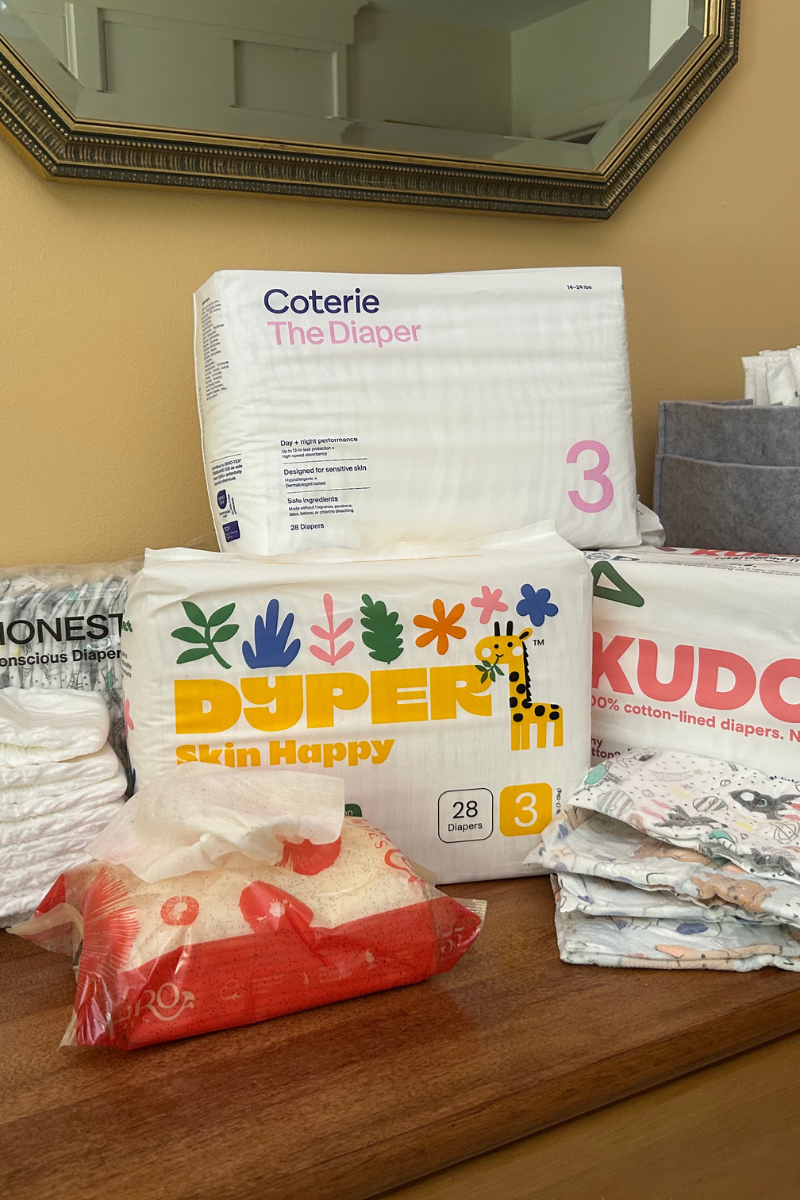 Various brands of size 3 diapers, including Coterie, Dyper, Kudos, and Honest, are displayed on a wooden surface with a mirror and other household items in the background. A red package is also visible in the foreground.