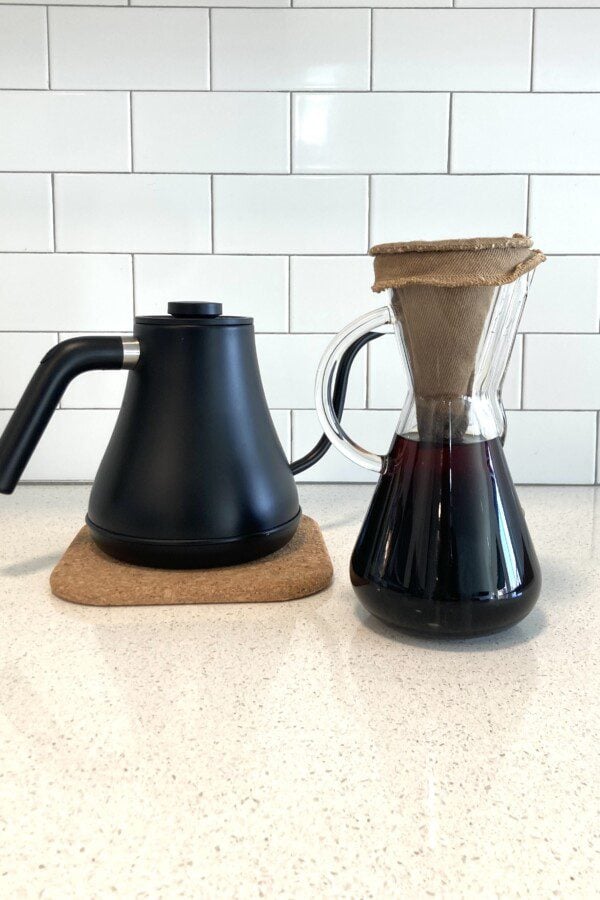 The photo shows a matte black gooseneck kettle on a cork coaster next to a glass carafe filled with coffee and topped with a cloth filter.