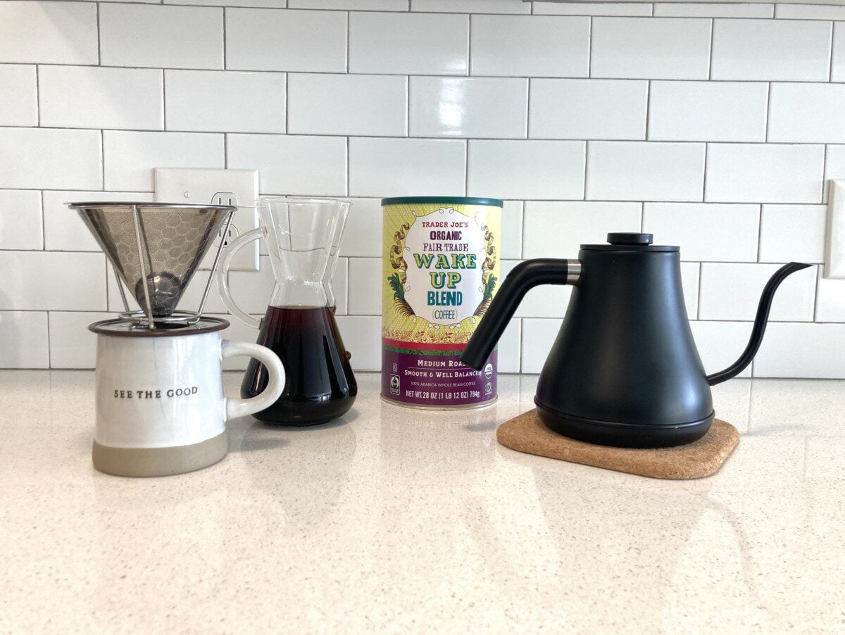 The photo features an array of coffee brewing equipment on a white countertop, including a matte black gooseneck kettle on a cork coaster, a canister of Trader Joe's "Wake Up Blend" coffee, a glass carafe, and a metal pour-over dripper resting on a mug.