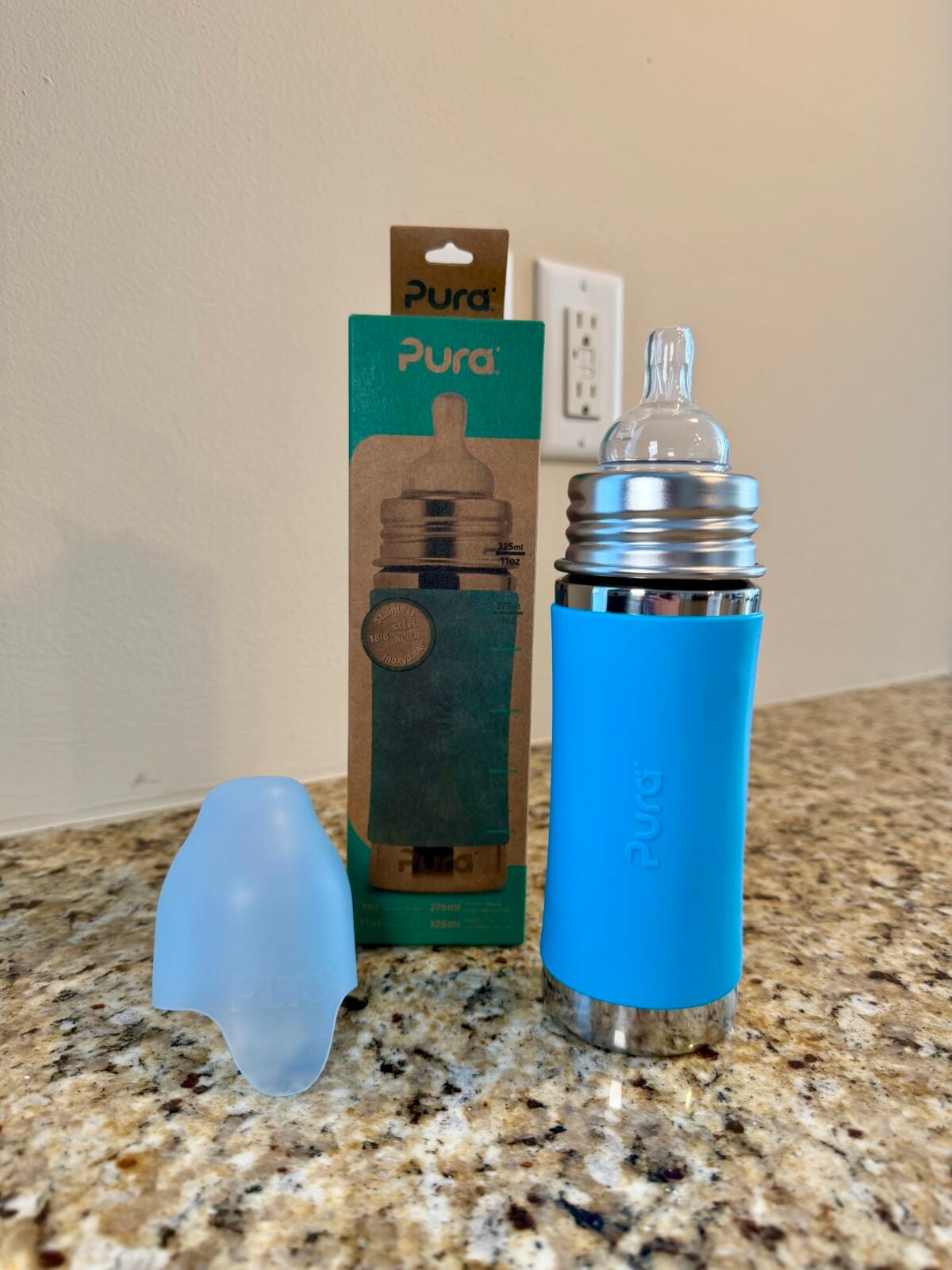 A stainless steel baby bottle with a blue silicone sleeve and a clear nipple is on a granite countertop. Next to it are a translucent bottle cap and the products cardboard packaging. The background shows a white wall and outlet.