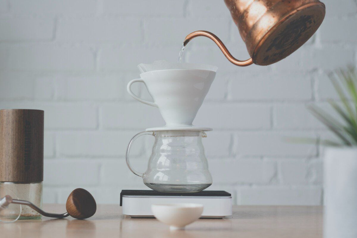 The photo shows hot water being poured from a copper gooseneck kettle into a white ceramic dripper as part of a pour-over coffee setup on a wooden table.