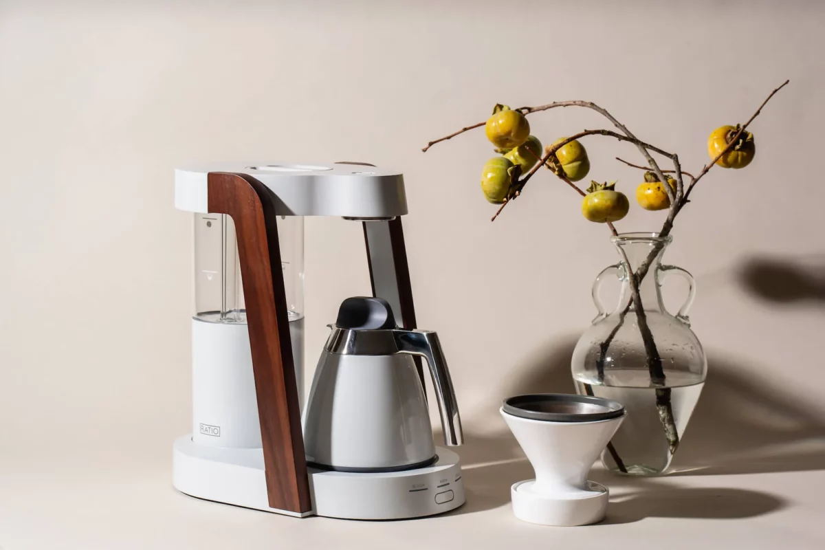 The photo features a sleek white and wood-trimmed Ratio coffee maker and a matching dripper base alongside a glass vase containing a branch with persimmons.