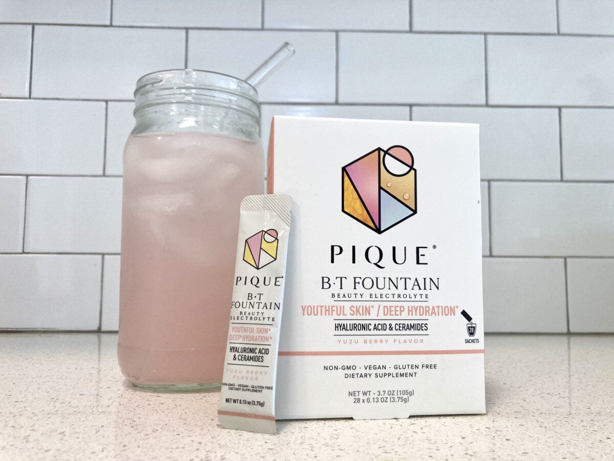 A glass of pink, prepared electrolyte drink sits next to a box and an individual sachet of Pique B-T Fountain Beauty Electrolyte in Yuzu Berry flavor.