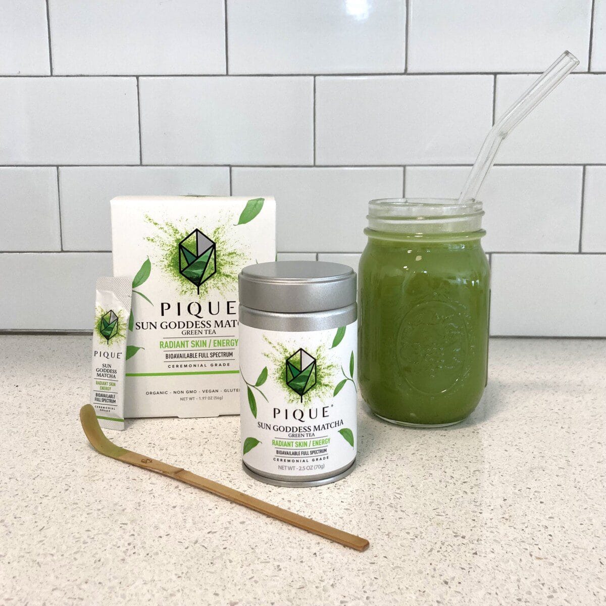 A Pique Sun Goddess Matcha kit is displayed on a white counter, featuring a box of tea packets, a canister of matcha, a bamboo whisk, and a glass mason jar with a glass straw filled with prepared green matcha.