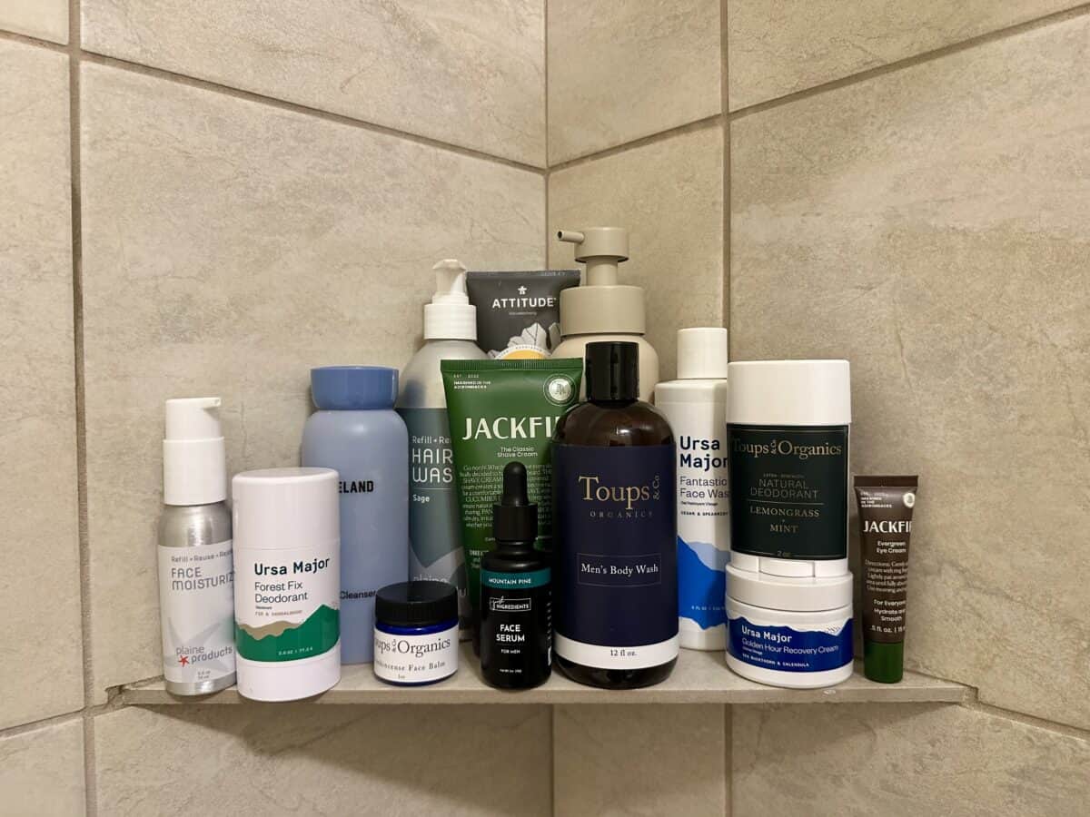 A shower shelf is stocked with various men's skincare products, including body wash, deodorant, face wash, face serum, hair wash, and other toiletries from brands like Ursa Major, Toups & Co. Organics, and Jackfir.
