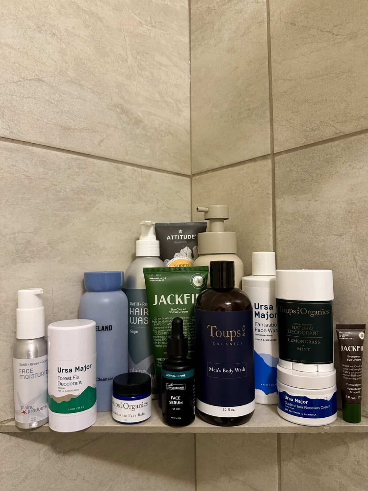 A shower shelf is stocked with various men's skincare products, including body wash, deodorant, face wash, face serum, hair wash, and other toiletries from brands like Ursa Major, Toups & Co. Organics, and Jackfir.