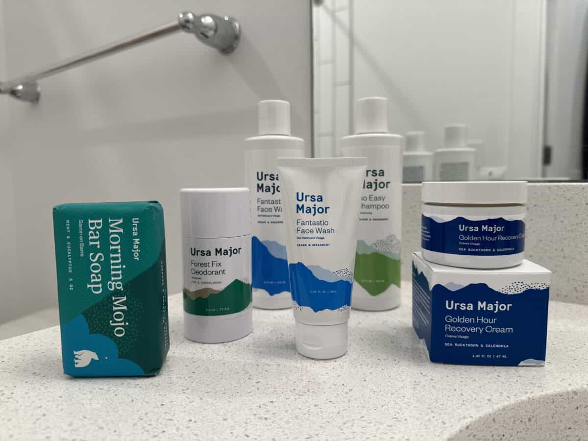 A collection of Ursa Major grooming products&mdash;including bar soap, deodorant, face wash, shampoo, and Golden Hour Recovery Cream&mdash;is arranged on a white bathroom counter against a mirrored background.