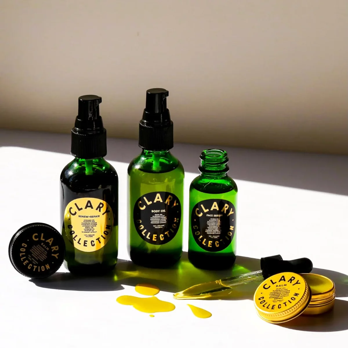 The photo displays three bottles of green-glass CLARY COLLECTION products&mdash;Renew+Repair Oil, Body Oil, and Face Serum&mdash;with yellow labels and three small round containers, one open with a dropper and yellow oil spilled on the white surface.