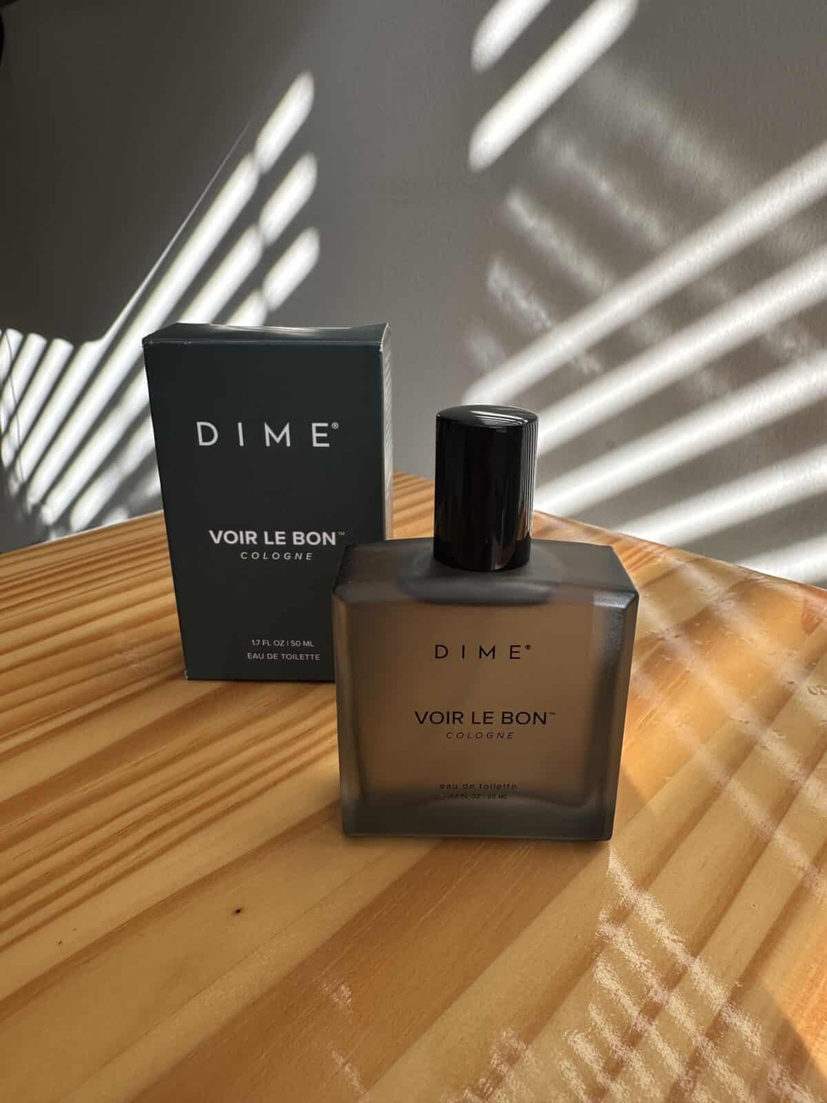 The photo shows a bottle of DIME "Voir Le Bon" cologne next to its box, sitting on a wooden surface with striped window shadows cast over it.