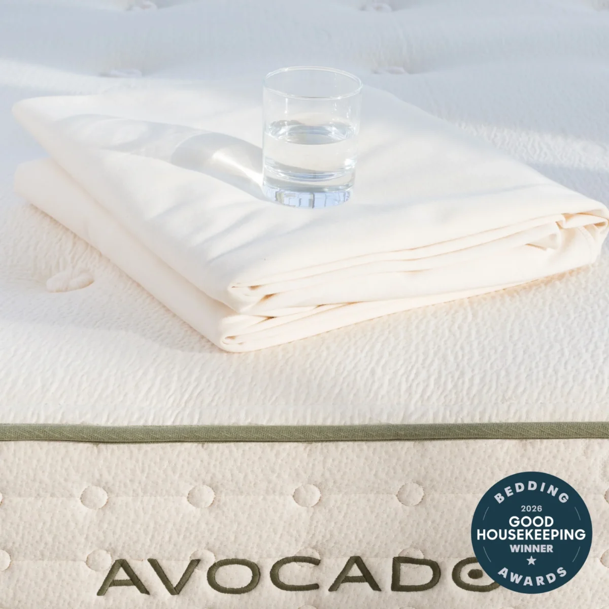 A folded white sheet and a glass of water sit on top of an Avocado mattress. Sunlight is shining on the scene. A Good Housekeeping 2020 Bedding Awards Winner badge is displayed in the lower right corner.