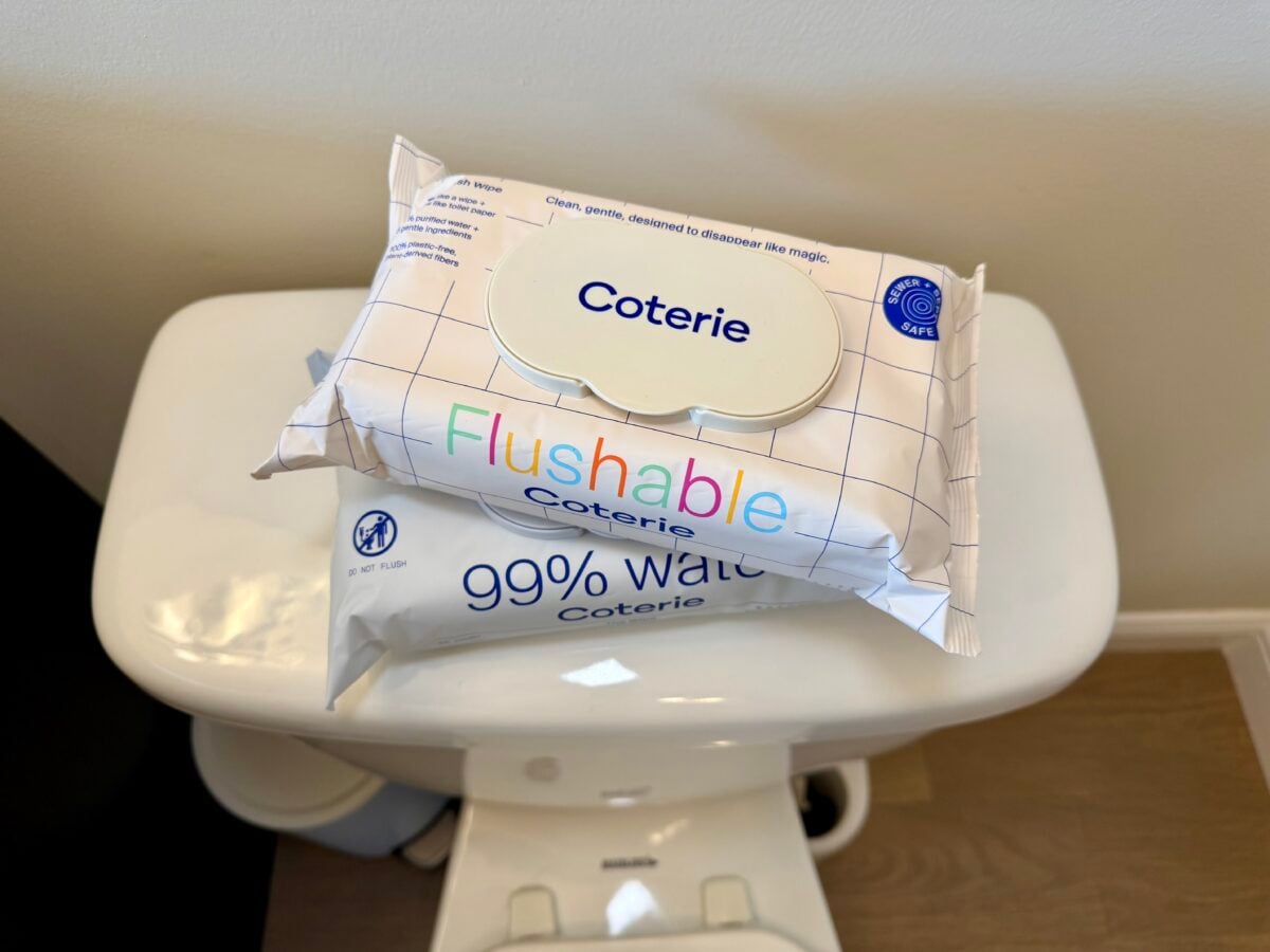 Two packs of Coterie wipes, one labeled Flushable and the other 99% Water, are stacked on top of a white toilet tank in a bathroom with light-colored walls and flooring.