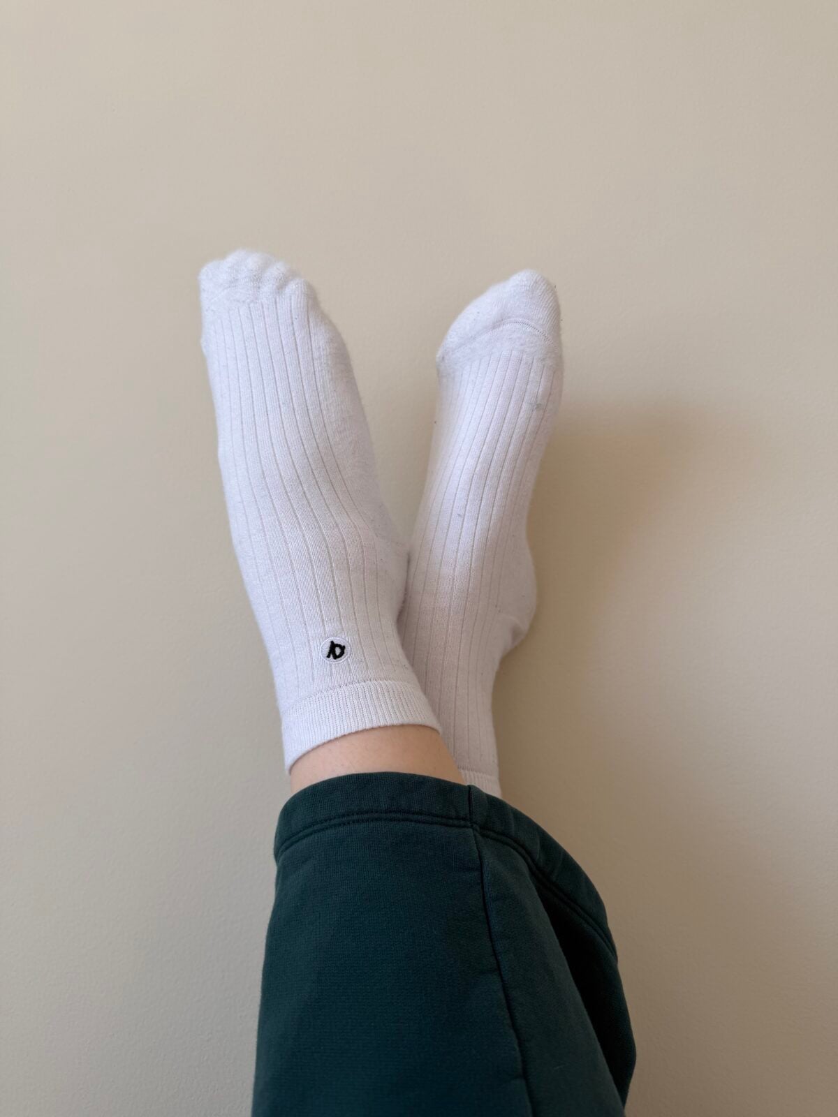 A person wearing white ribbed socks with a small black logo rests their crossed feet against a plain light-colored wall, dressed in dark green pants.